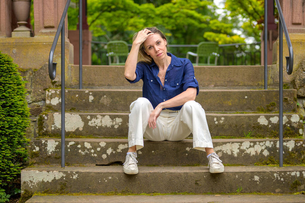 Egy kék inges és fehér nadrágos anya elmélkedve ül a parkban a lépcsőn, és a haját simogatja.. - Fotó, kép