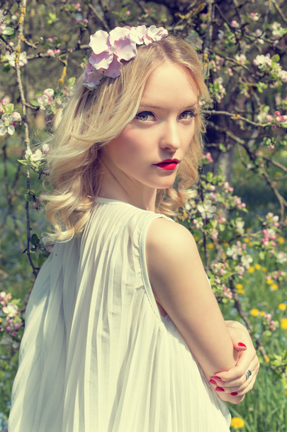 mooi offerte zoete jonge meisje met een krans van bloemen in haar haar in een witte lichte jurk wandelingen in de weelderige tuin - Foto, afbeelding