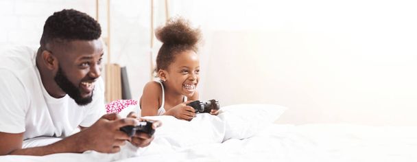 Czarny ojciec i córka leżą w łóżku i śmieją się, grając w gry wideo. Ojciec ma na sobie białą koszulkę, a córka ma na sobie różowo-białą koszulę, przestrzeń do kopiowania. - Zdjęcie, obraz