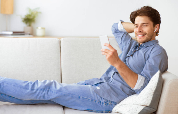 Hombre en jeans y camisa azul se relaja en un sofá blanco con teléfono y auriculares, apoyado hacia atrás con expresión relajada. - Foto, Imagen