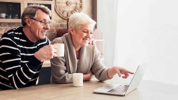 Egy idősebb pár ül egy asztalnál az otthonukban. A nő a laptop képernyőjére mutat, miközben a férfi egy bögre kávét tart a kezében, és őt nézi.. - Fotó, kép