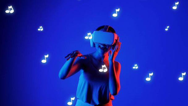 Mujer sonriente escuchando y bailando en canción divertida usando auriculares VR en nota gráfica melodía musical moderno diseño en estudio iluminación dinámica en música y tecnología concepto fondo azul. Alucinaciones. - Foto, Imagen