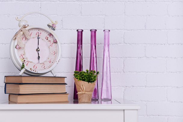 Interior design with alarm clock, plant, decorative vases and stack of books on tabletop on white brick wall background - 写真・画像