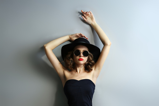 Portrait of beautiful model in black dress, hat and sunglasses on gray background - Foto, afbeelding