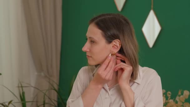 Μια γυναίκα προσαρμόζει το σκουλαρίκι της ενώ στέκεται σε ένα κομψό δωμάτιο με πράσινους τοίχους και γεωμετρικούς καθρέφτες. - Πλάνα, βίντεο