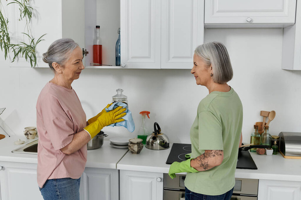 Een volwassen lesbisch echtpaar reinigt samen hun moderne appartementenkeuken. Een vrouw draagt gele handschoenen en veegt een glazen pot af.. - Foto, afbeelding