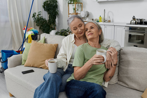 A mature lesbian couple relaxes on a couch in their modern apartment after cleaning, enjoying a cup of coffee. - Photo, Image