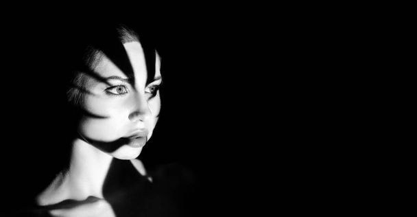 Černobílý portrét ženy s tváří částečně zakrytou stínovým, světlým vzorem. Její oči jsou viditelné a dívají se nalevo, dávají obraz pocit tajemství a intrik. - Fotografie, Obrázek