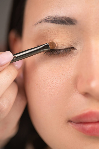 Extreme close-up van het ooglid van een vrouw met een make-up borstel aan te brengen glinsterende oogschaduw. Het detail benadrukt de textuur van de huid en de precisie van de oogschaduwapplicatie, met de nadruk op schoonheid - Foto, afbeelding