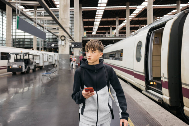 Teenager walking on a train platform in a big city, holding a phone and looking into the distance. - Photo, Image