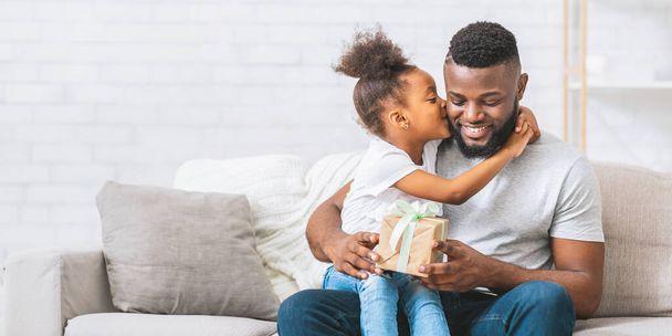 Afrikkalainen amerikkalainen nuori tyttö, jolla on tummat hiukset, istuu vaalealla sohvalla ja syleilee isäänsä antaen hänelle lahjan. Isä hymyilee katsoessaan lahjaa.. - Valokuva, kuva