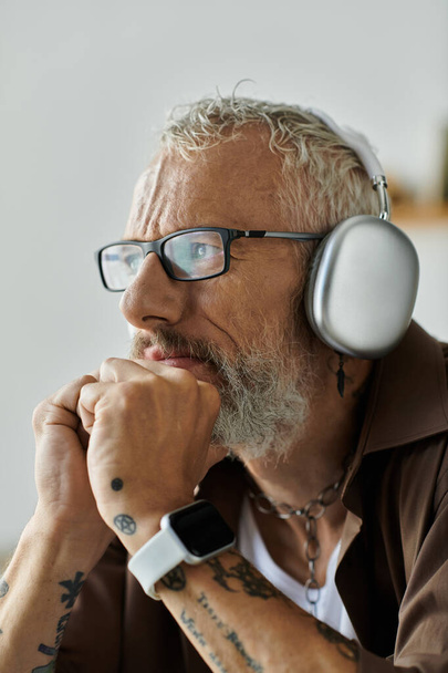 A mature gay man with gray hair and tattoos works remotely from home while wearing headphones. - Photo, Image