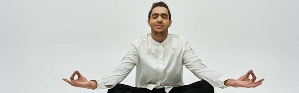 Un homme afro-américain élégant est assis dans une posture méditative, portant une chemise blanche et un pantalon noir sur un fond gris. - Photo, image