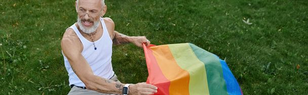 Un homme gay mature avec des tatouages et une barbe grise tient un drapeau arc-en-ciel à l'extérieur. - Photo, image
