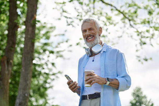 Ein reifer schwuler Mann mit Tätowierungen und grauem Bart genießt einen Kaffee und einen sonnigen Tag im Park. - Foto, Bild
