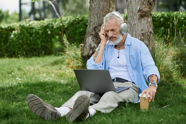 Egy érett, tetovált, szürke szakállú meleg férfi ül a szabadban a füvön, zenét hallgat fejhallgatón, miközben laptopot használ.. - Fotó, kép