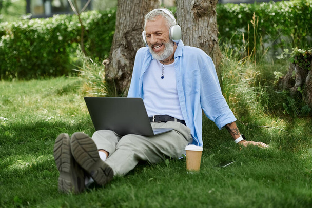 Een volwassen gay man met tatoeages en een grijze baard ontspant in een park, luisteren naar muziek en werken aan zijn laptop. - Foto, afbeelding