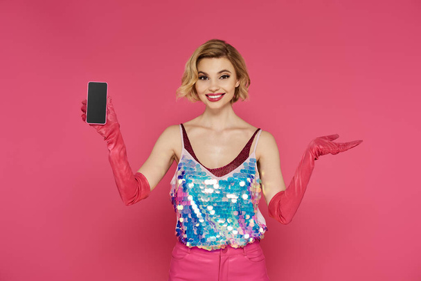 Mujer en rosa lentejuelas sonrisas superiores con smartphone, telón de fondo rosa. - Foto, imagen