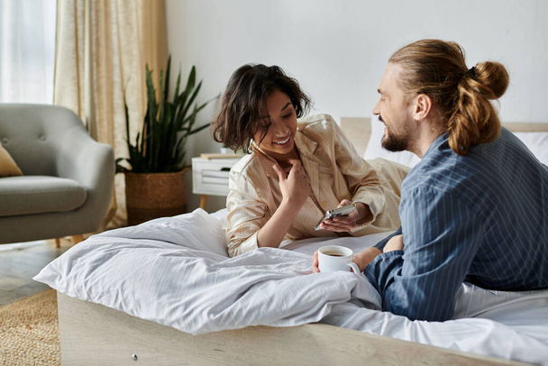 Um casal abraça-se em uma cama, desfrutando de uma xícara de café e um momento de riso juntos. - Foto, Imagem