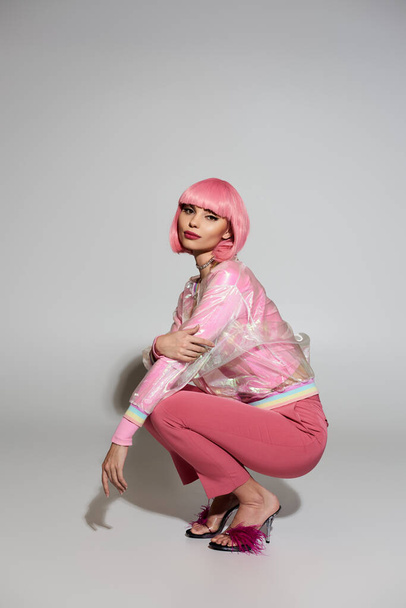 Una mujer con el pelo rosa se sienta en una postura en cuclillas con una chaqueta rosa transparente y pantalones a juego. - Foto, Imagen