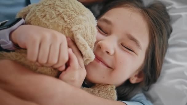 Morning kid embracing toy bonding to unknown mother at bed closeup. Small funny child playing favourite teddy bear spending time with unrecognizable mom. Toddler girl awaking at soft pillows relaxing - Felvétel, videó