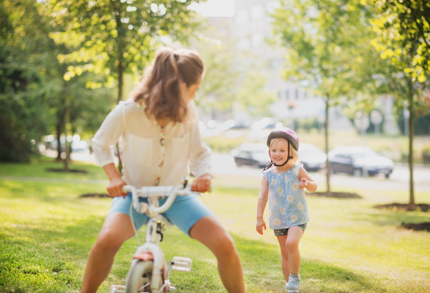 Mother playfully riding a blonde girl's bike in a sunny park - Φωτογραφία, εικόνα