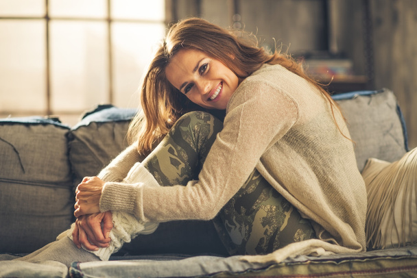 Smiling woman on sofa in loft, hugging her knees - Fotó, kép