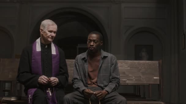 Starší kněz v sutaně sedí na lavičce vedle mladého černocha drží růženec korálky a mluví s ním v kostele - Záběry, video