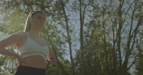 Een zelfverzekerde jonge vrouw met handen op haar heupen rust van haar training buiten onder de zon, die de begrippen fitness, vastberadenheid en welzijn belichaamt. - Video