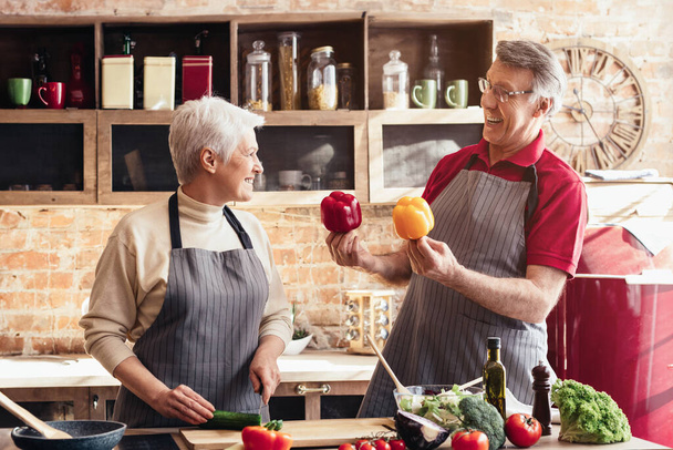 Un couple de personnes âgées se tient ensemble dans une cuisine familiale, préparant un repas. La femme coupe des légumes sur une planche à découper, tandis que l'homme regarde avec un sourire. Ils portent tous les deux des tabliers - Photo, image