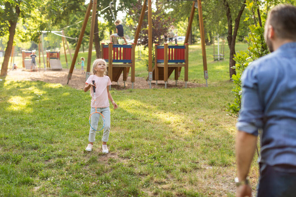 Ένα νεαρό κορίτσι με ξανθά μαλλιά παίζει με ένα σχοινάκι σε ένα πάρκο. Φοράει ροζ πουκάμισο, μπλε τζιν και λευκά αθλητικά παπούτσια. Πίσω της είναι μια ξύλινη παιδική χαρά με κόκκινη στέγη. - Φωτογραφία, εικόνα