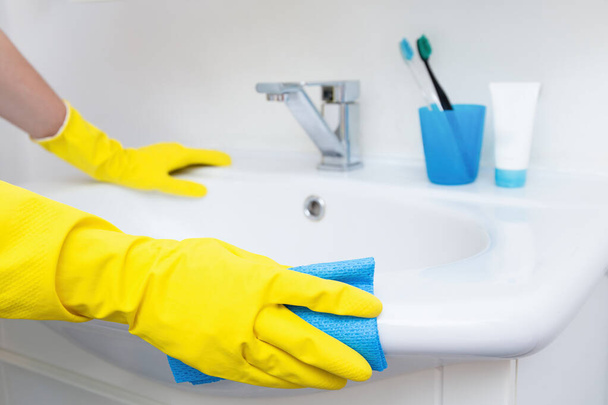 Gele handschoenen reinigt een badkamer wastafel met een blauwe doek, huishoudelijke hygiëne - Foto, afbeelding
