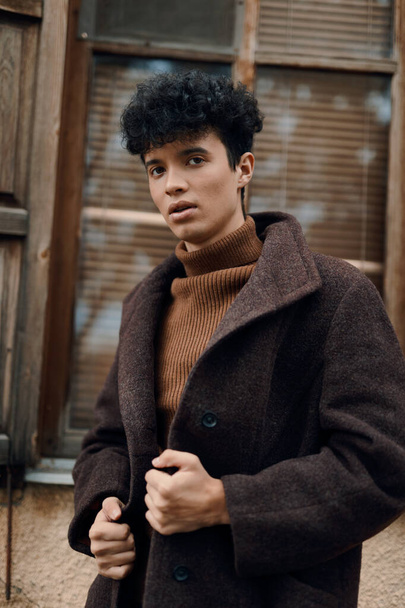 Jongeman met krullend haar in bruine jas en schildpadden nek trui voor het raam met houten luiken. mode stijl. - Foto, afbeelding