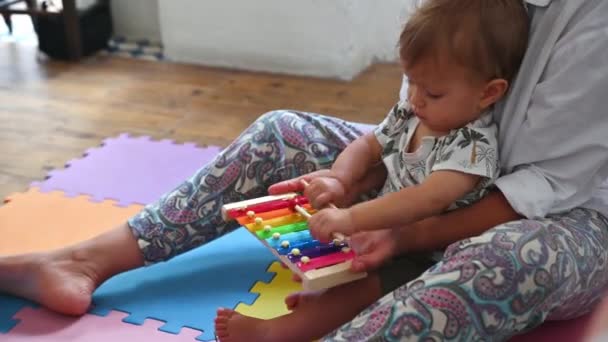 Adorable smart baby boy is playing a colorful xylophone under the guidance of his mother. A 10-month-old Caucasian child knocks wooden sticks on multicolored iron plates, studying the rhythm of music - Imágenes, Vídeo