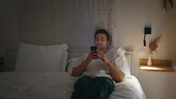 Sleepless man browsing phone in bedroom lit by soft lamp light. Relaxed guy using smartphone watching social media content in bed dark apartment. Calm male looking cellphone screen relaxing at bedtime - Footage, Video