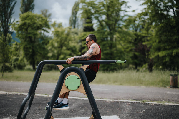 Ein fitter, tätowierter Mann trainiert mit horizontalen Metallstangen in einem Stadtpark, der von Grün und Bäumen umgeben ist. - Foto, Bild