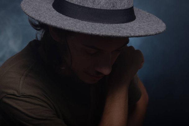 Close-up portrait of thoughtful young man wearing gray colored hat. Isolated on smoky black background. - Zdjęcie, obraz