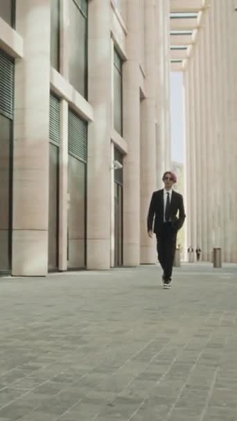 Pionowe dolly-in pełne powoli elegancki azjatycki różowłosy mężczyzna model w formalnym garniturze i okulary przeciwsłoneczne przechodząc przez kamerę z ufnością na zewnątrz - Materiał filmowy, wideo