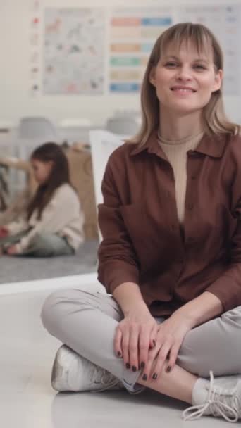 Κάθετο πορτρέτο του χαμογελαστού Καυκάσου εκπαιδευτή γυναίκα που εργάζεται στο νηπιαγωγείο κάθεται στο πάτωμα με τα πόδια σταυρωμένα και με αυτοπεποίθηση κοιτάζοντας κάμερα - Πλάνα, βίντεο