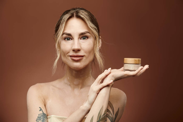 A young woman with vitiligo and a tattoo holds a cream jar in her hand, smiling confidently against a beige background. - Foto, immagini
