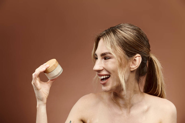 A young woman with vitiligo and a tattoo smiles while holding a jar of cream against a beige background. - Foto, immagini