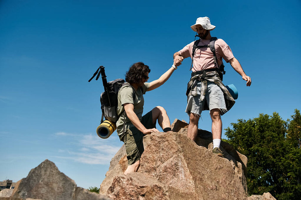 Gay couple hiking together on rocky path in wilderness, both with backpacks, enjoying summer weather. - Photo, Image