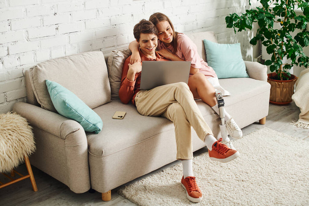 Ένα νεαρό ζευγάρι κάθεται σε έναν καναπέ, κοιτάζοντας ένα φορητό υπολογιστή μαζί. - Φωτογραφία, εικόνα