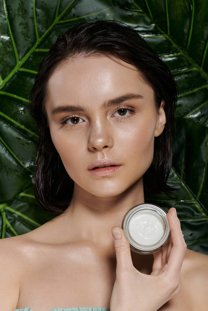 A woman with clear skin holds a cream jar in front of lush green foliage. - Photo, image