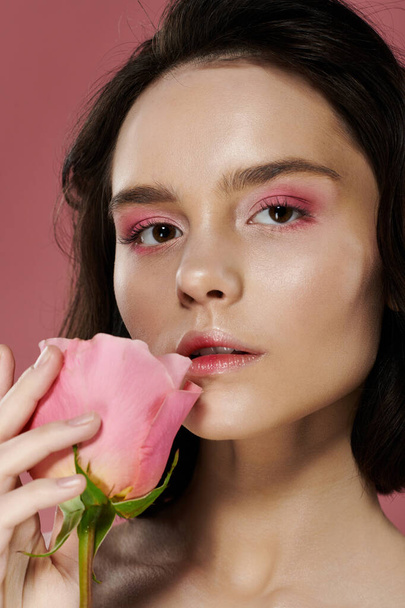 Een vrouw met roze oogschaduw houdt een zachte roze roos tegen haar lippen. - Foto, afbeelding
