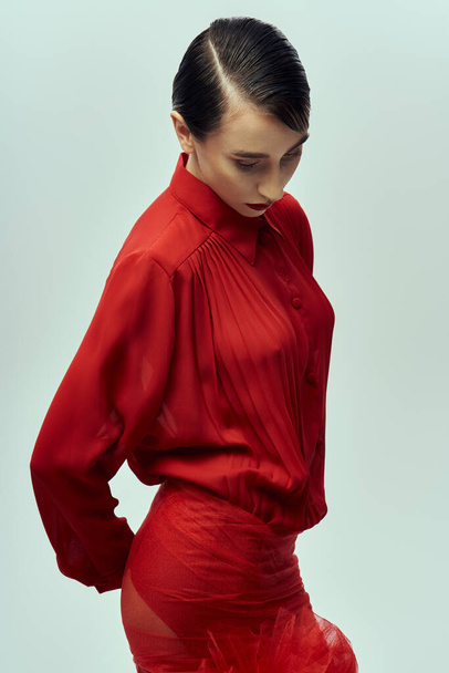 Una mujer joven con el pelo negro corto posa en un vestido rojo que fluye, exudando confianza y estilo sobre un fondo gris. - Foto, Imagen