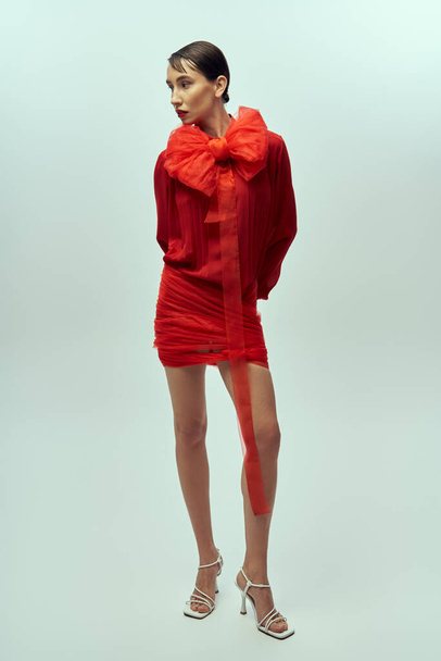 Egy fiatal nő piros ruhában, nagy masnival, szürke háttérrel pózol.. - Fotó, kép