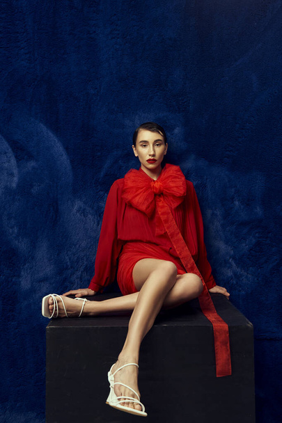 A young woman with short hair poses in a red outfit with a large bow against a blue background. - Fotografie, Obrázek