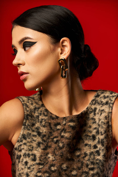 Dark haired woman with bold makeup poses against red backdrop in leopard-print dress. - Photo, Image