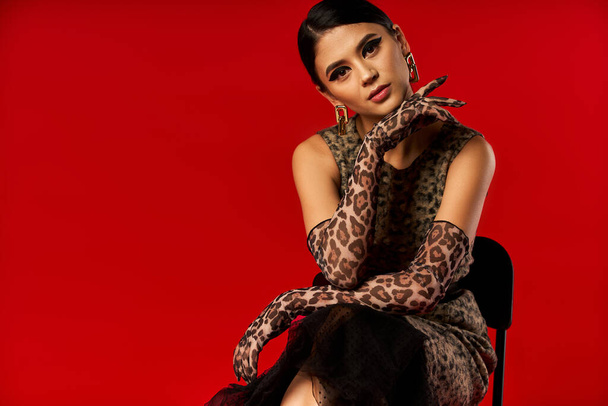 A young woman wearing a leopard print dress and gloves poses against a bright red background. - Foto, Imagem